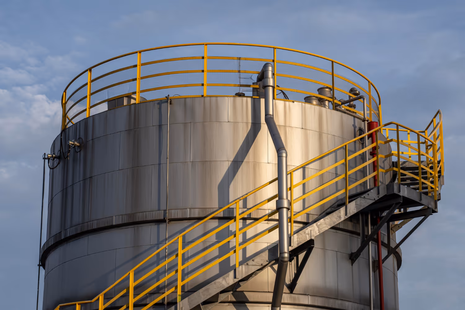 Close-up of a large industrial metal storage tank with yellow handrails and an exterior staircase against a blue sky.