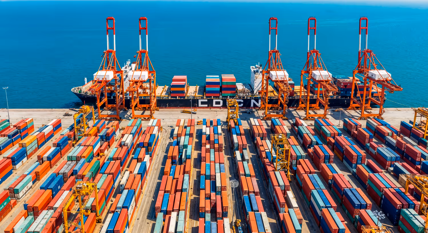 Aerial view of a cargo ship docked at a port with multiple colorful shipping containers and large orange cranes unloading the containers.