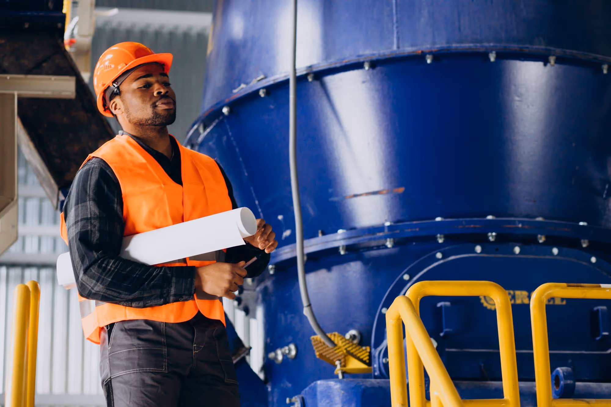Industrial worker wearing an orange helmet and safety vest holding rolled blueprints inside a factory with large blue machinery.