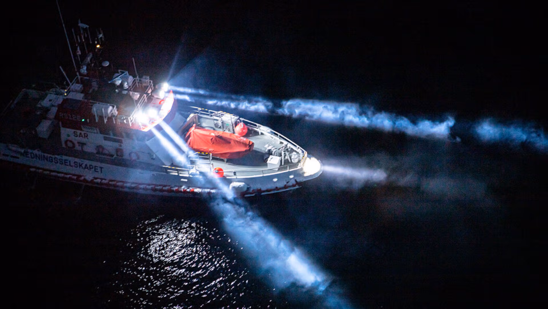 Rescue boat shining multiple powerful searchlights across dark water at night.
