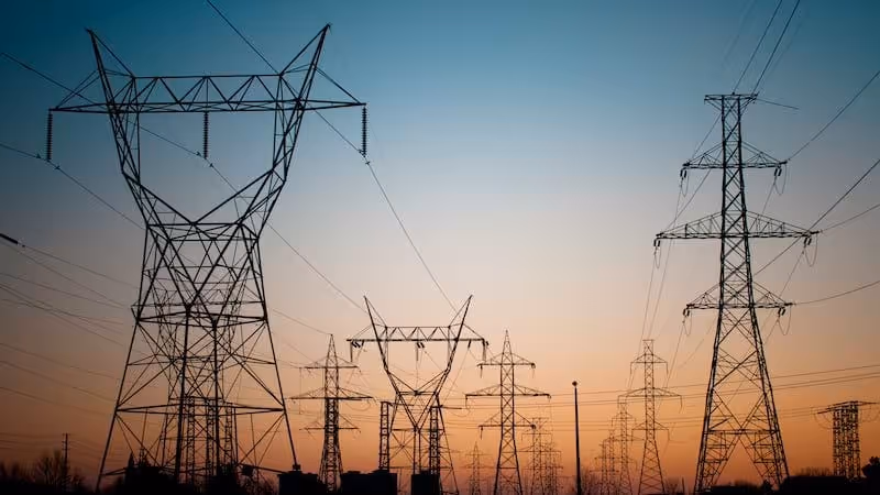Silhouettes of multiple high voltage power lines and towers against a sunset sky.